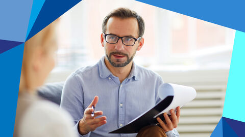 Ein mittelalter Mann mit Bart und Brille hält Unterlagen in der Hand und einen Stift, er gestikuliert. Er sitzt einer Frau gegenüber, die man in Unschärfe sieht.