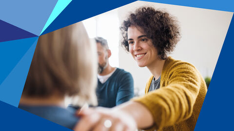 Foto zeigt eine freundlich lächelnde Frau, die ihre Hand auf die Schulter einer ihr zugewandten Person legt.