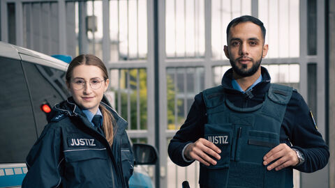 Eine junge Justizvollzugsbeamtin und ein junger Justizvollzugsbeamter Stehen vor einen Justizfahrzeug.