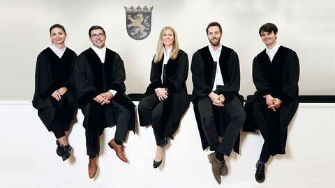 Fünf junge Menschen in Roben sitzen vor einem weißen Hintergrund.